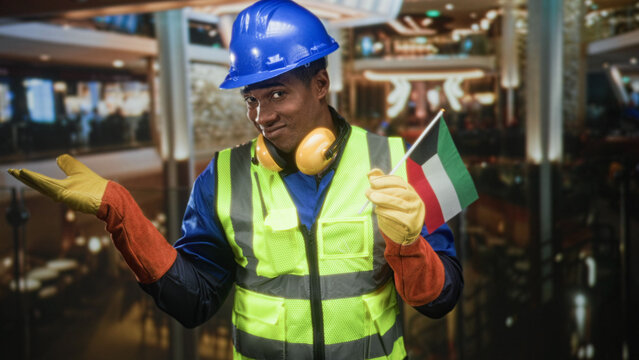 Man construction worker in hardhat holds kuwait flag and gestures palm up in hotel lobby; pride solidarity.