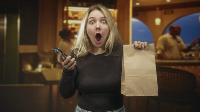 Woman holding smartphone and brown paper delivery bag, mouth open and raising bag while looking at phone in building lobby; surprise delivery issue.