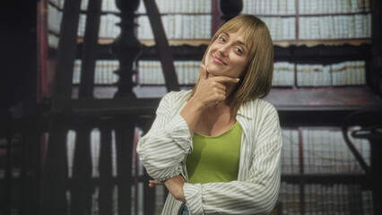 Fototapeta premium Woman with hand on chin thinking in a library, middle age blonde wearing green tank and striped shirt, slight smile and folded arm; quiet contemplation.