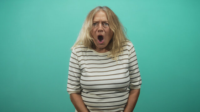 Blonde senior woman with open mouth and hands clasped in front, striped shirt in a studio teal backdrop; surprise playfulness.