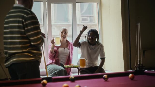 Young adult man pocketing ball with precise shot then fist bumping cheering friends while playing billiards at bar over beers