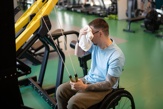Strong focused disabled man in wheelchair wiping sweat with towel while finishing workout on gym equipment at rehabilitation training. Male with legs immobility taking break during powerful exercises.