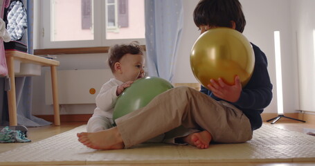 Fototapeta premium Baby and young boy playing together on floor with golden and green balloons in cozy bedroom, joyful sibling interaction and tender family bonding moment