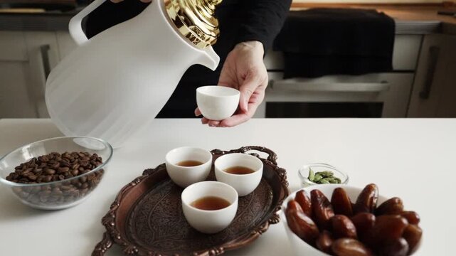 close-up of arabic coffee being poured from a white dallah into a small finjan cup on a copper tray with dates, coffee beans and cardamom nearby