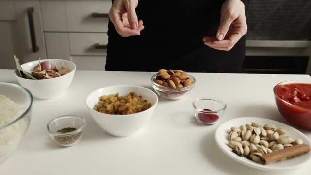 close-up of hands holding a glass bowl of almonds beside garlic raisins pistachios tomato sauce and spices, showing classic ingredients for a rich middle eastern style rice dish
