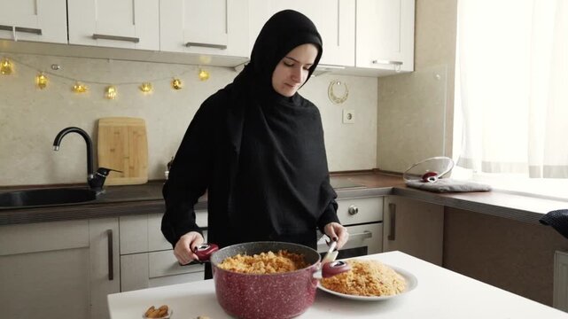 muslim woman in black hijab transfers fluffy tomato colored rice from a red cooking pot onto a large serving platter, assembling a hearty family style main course in the kitchen
