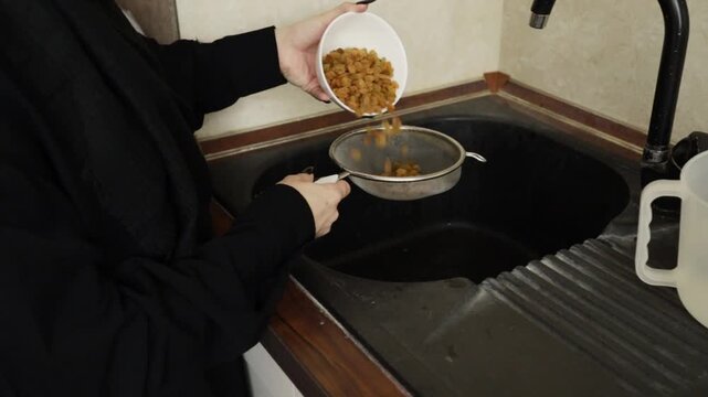 hands tip a white bowl of golden raisins into a metal sieve above the sink, capturing the transfer step before washing and draining ingredients for a savory rice recipe