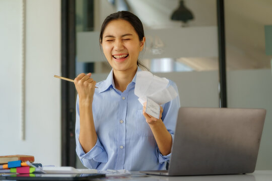 Happy Asian businesswoman celebrating financial success holding receipts at desk with laptop.