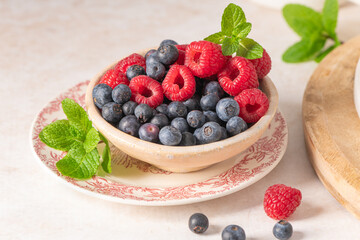 A vibrant small bowl filled with fresh red raspberries and blueberries garnished with green mint sprigs on a decorative plate with scattered berries and a wooden board in a bright kitchen © anammarques