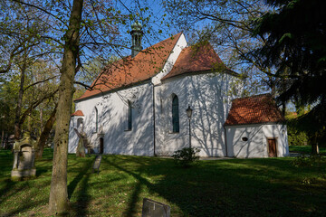 Fototapeta premium Pisek, Czech Republic - April 18, 2026 - Memorial park at the Holy Trinity. Historic white church with a red tiled roof surrounded by trees on a sunny day