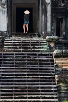 A beautiful Southeast Asian woman inspects a historical structure wearing a conical hat and a denim miniskirt at the temple gate - Ancient temple complex Angkor Wat garden - Siem Reap, Cambodia