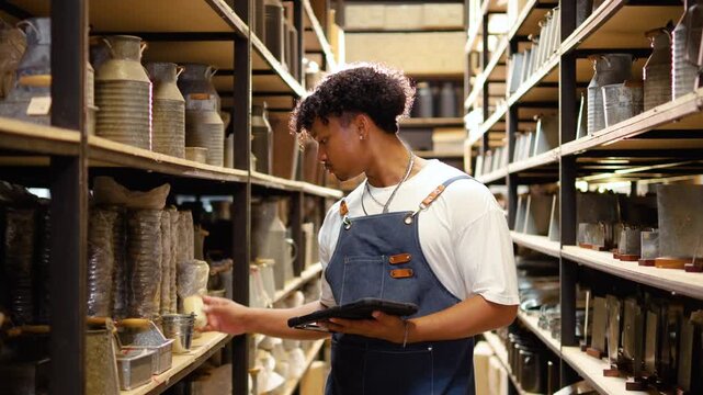 Focused young Black man in a denim apron uses a tablet for inventory management in a pottery warehouse. He checks ceramic stock on shelves, representing small business retail and logistics work.