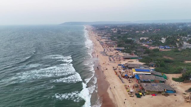 Drone shot gliding over Baga Beach, showcasing a long sandy coastline filled with beach activity, colorful seating, and rhythmic ocean waves, blending natural beauty with energetic coastal life.