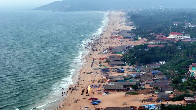 Cinematic aerial view of Baga Beach packed with beachgoers, colorful umbrellas, and lively shacks, as waves crash along the shoreline creating a vibrant and energetic coastal scene.
