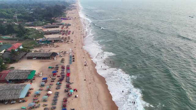 Cinematic aerial view of Goa&rsquo;s vibrant coastline, with rows of colorful beach umbrellas, lively shacks, and waves rolling onto the sandy shore. The long stretch of beach captures energy and coastlines