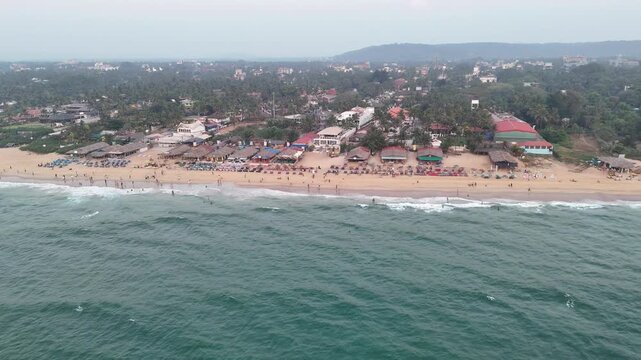 Drone shot capturing a bustling coastline with beachside caf&eacute;s, neatly arranged sunbeds, and soft waves rolling in. The scene blends relaxed seaside life with greenery and distant hills.