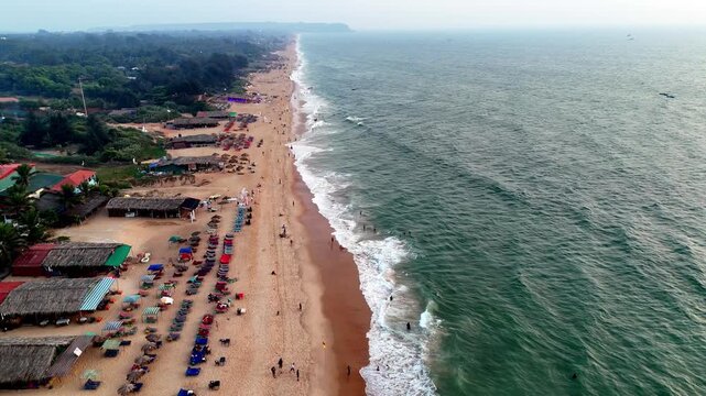 Wide aerial perspective of a vibrant beach featuring rows of umbrellas, active shoreline, and lush palm-covered surroundings. The ocean&rsquo;s edge contrasts beautifully with the lively coastal landscape.