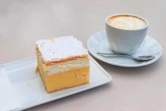 The original Bled cream cake (Cremeschnitte) on white plate and cup of cappuccino on a table. Traditional Slovenian dessert