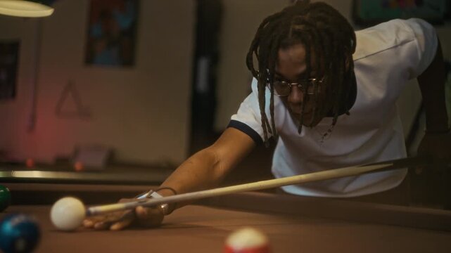 Focused young adult Black man with dreadlocks aiming cue at pool ball then striking precise shot during billiards game at club, slow motion