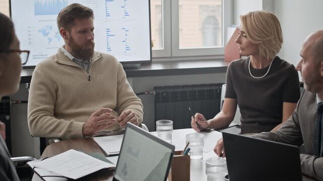 Medium shot of four colleagues sitting at desk and discussing business strategy, examining documents and schemes
