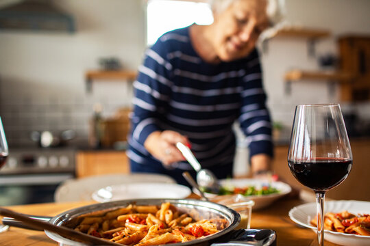 Older woman serving pasta and wine in cozy home kitchen