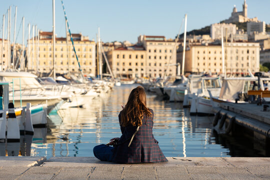 Femme vue de dos avec cheveux longs bruns assise sur le quai du Vieux-Port de Marseille, regardant les bateaux et la colline avec la basilique Notre-Dame de la Garde, lumi&egrave;re douce et chaude