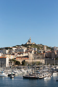 Vue sur le Vieux-Port de Marseille avec des bateaux &agrave; quai au premier plan et la basilique Notre-Dame de la Garde sur la colline en arri&egrave;re plan, b&acirc;timents anciens et ciel bleu, format vertical