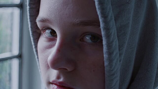 A sad teenage girl wearing a gray hoodie. She has a serious expression, reflecting feelings of loneliness and stress. Soft natural light illuminates her face.