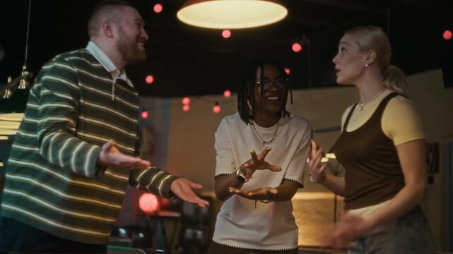 Medium full shot of cheerful young adult woman losing break shot in billiards game while playing rock paper scissors with two male friends at pool hall