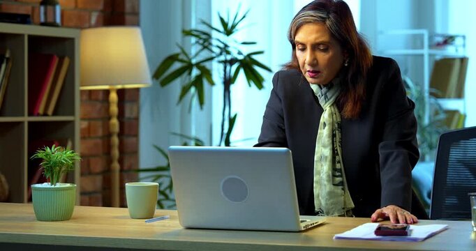 Senior Indian businesswoman in corporate attire working while leaning on laptop standing near desk in modern cozy office space experienced female executive with white hair in a professional setting