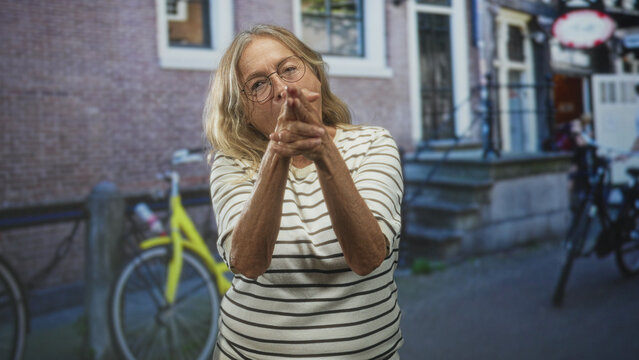 Senior woman points fingers forward in a finger gun gesture on a street beside bicycle and brick building, wearing striped shirt and glasses; playful gesture.