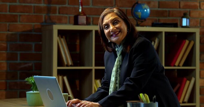 Senior Indian businesswoman in corporate attire working on laptop in cozy office space looks at camera with a smile then poses with folded hands and a thumbs up experienced female executive