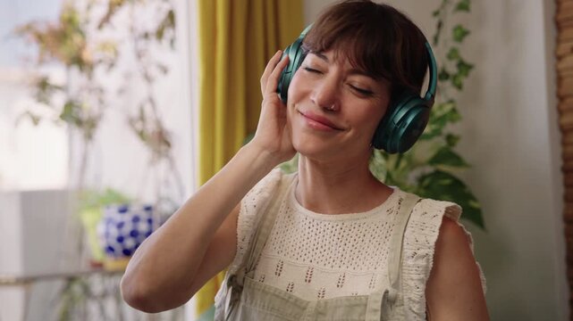 Woman enjoying music with headphones at home, eyes closed and smiling. Relaxation, wellbeing and self care in an authentic and cozy everyday lifestyle moment.