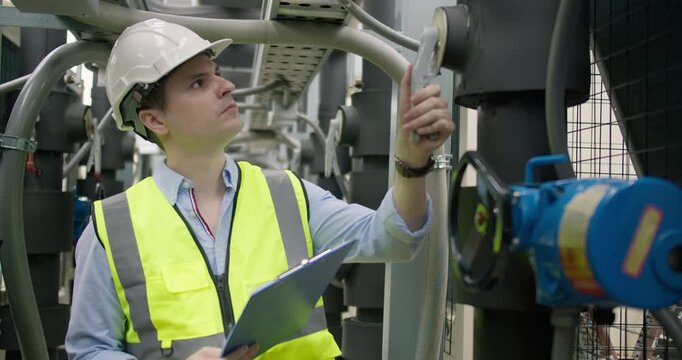A male engineer performs a maintenance check on an industrial piping system, using a tool and clipboard to ensure operational safety and efficiency.