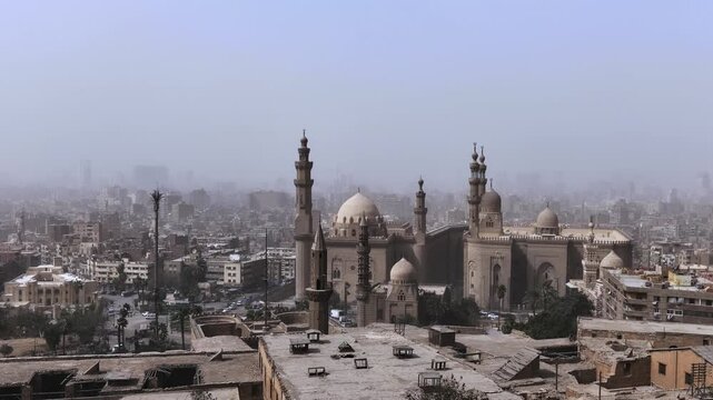 Panoramic view of Sultan Hassan and Al-Rifa'i mosques rising above dusty Cairo skyline, Egypt, with towering minarets and massive stone domes in medieval Islamic architecture.