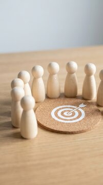 A group of wooden peg figures are arranged around a cork target with an arrow in the bullseye, representing teamwork, focus, and achieving objectives.
