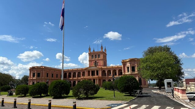 Pal&aacute;cio de L&oacute;pez e bandeira do Paraguai sob c&eacute;u azul em Assun&ccedil;&atilde;o. Sede do governo nacional, arquitetura neocl&aacute;ssica rosada e jardins oficiais da capital paraguaia.