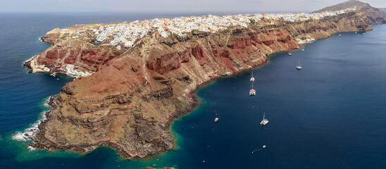 A wide-angle drone panorama of Oia, Santorini, featuring the whitewashed village perched on immense red volcanic cliffs with catamarans anchored in the deep blue Aegean Sea. © bardhok