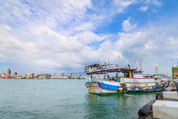 Obraz premium A colorful fishing boat moored at the pier in Kaohsiung Harbor, Taiwan, with the city skyline visible under a bright summer sky.