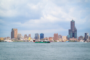Obraz premium A scenic view of the Kaohsiung harbor skyline with modern skyscrapers and a passenger ferry boat crossing the water on a summer afternoon in Taiwan.