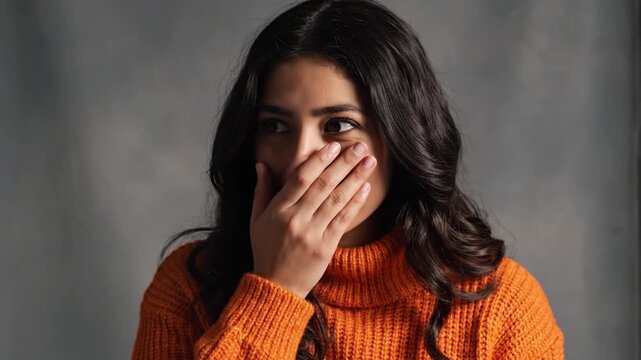 A woman in an orange top covers her mouth and shows different expressions. Her background is gray, and she appears engaged in the moment. The setting highlights her reactions