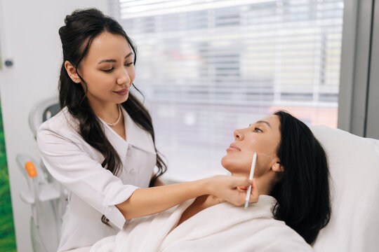 Portrait of woman cosmetologist talking with female client, using white pencil to mark lines on face before cosmetic procedure, preparing for plastic surgery or beauty injections.