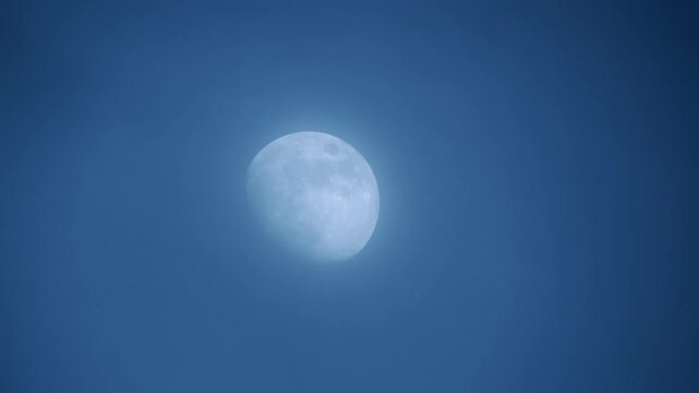The moon behind moving thin clouds in the blue sky. Astrophotography footage of the lunar view at dusk.