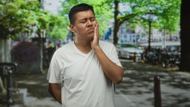 Hispanic man holding cheek with closed eyes and wince on street near parked bikes and lamppost; toothache pain.