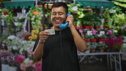 Fototapeta premium Man holds blue handset to ear and studies a white creditcard while smiling at an outdoor flower market; joy commerce.
