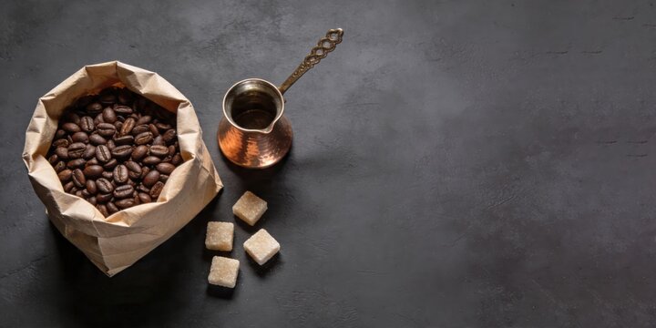 Bag of roasted coffee beans beside copper cezve and sugar cubes on dark textured surface. Traditional coffee brewing setup for morning ritual. banner format