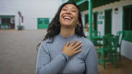 Woman laughing with hand covering mouth and other hand on chest in building courtyard, wearing hoop earrings and ribbed sweater  joyful surprise gratitude. © Krakenimages.com