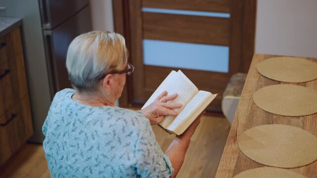 White grandma studying recipe at table, elderly white woman with glasses flips through cookbook over wooden table with round placemats, focused hands and pages, domestic kitchen scene, home cook