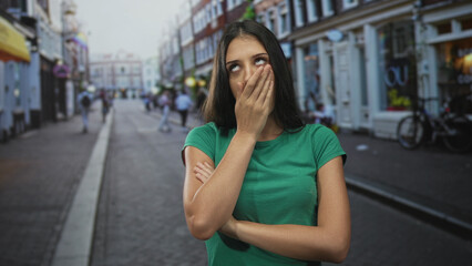 Fototapeta premium Young woman with arms crossed and hand over mouth on city street, eyes rolled and closed mouth; annoyance.
