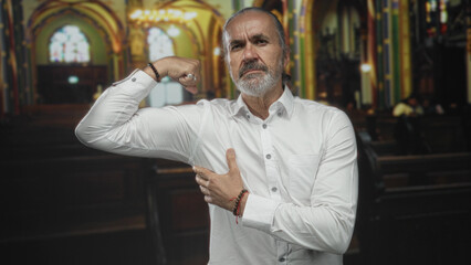 Obraz premium Man flexing bicep and pressing palm to chest inside ornate church building wearing white shirt and bracelets; faith confidence.